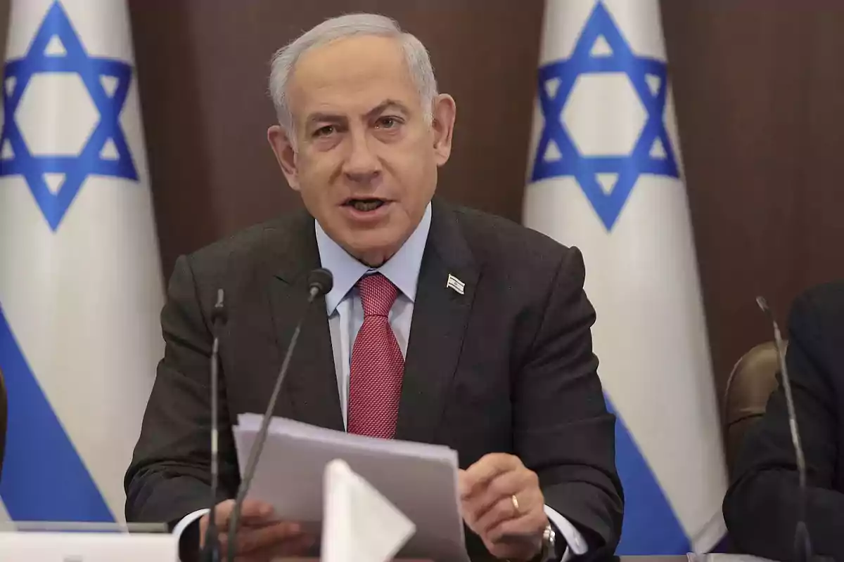 Gray-haired man in a dark suit and red tie speaking in front of microphones with Israeli flags in the background Gray-haired man in a dark suit and red tie speaking in front of microphones with Israeli flags in the background
