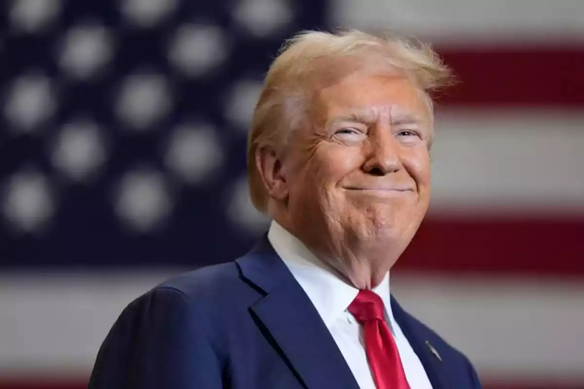 Hombre de cabello rubio con traje azul y corbata roja sonriendo frente a una bandera de Estados Unidos desenfocada