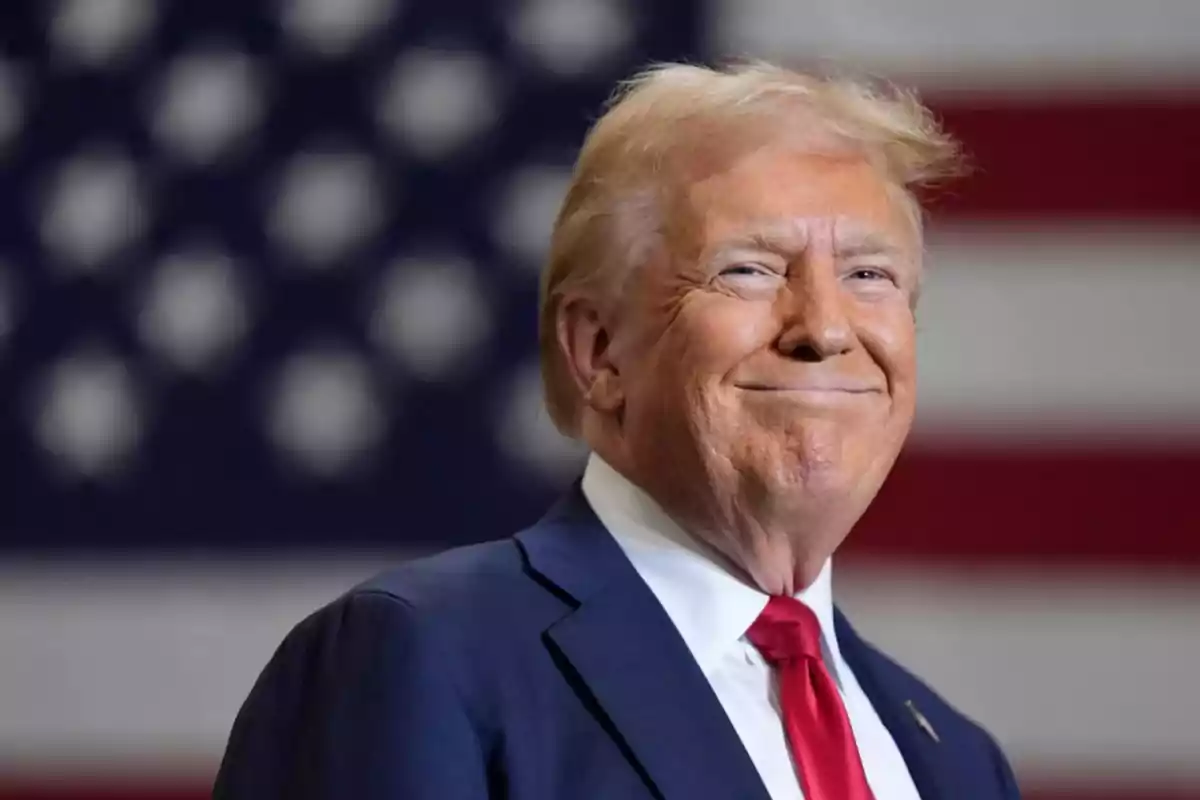 Blond-haired man in a blue suit and red tie smiling in front of a blurred United States flag