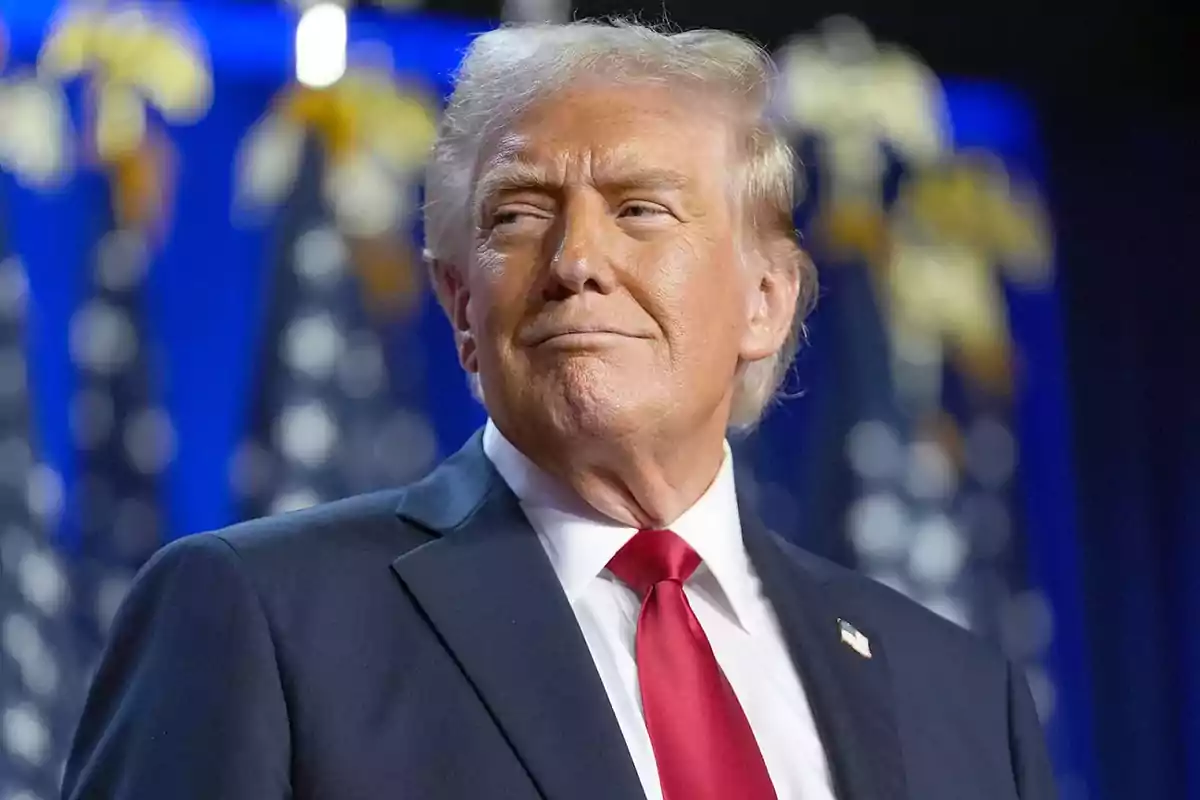 Hombre de cabello rubio con traje oscuro y corbata roja frente a un fondo de banderas estadounidenses desenfocadas