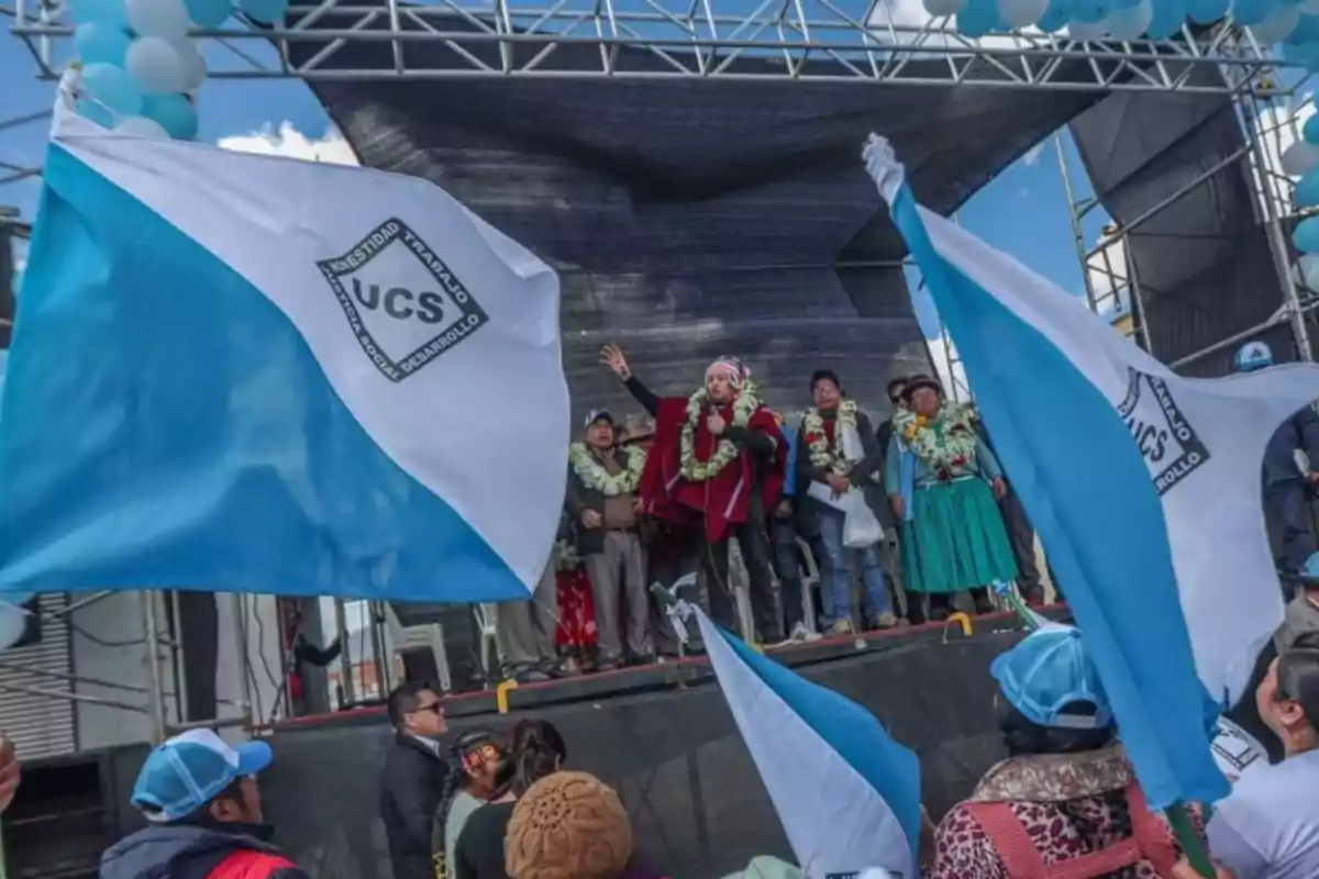 People on a stage during a political event with UCS flags waving and blue and white decorative balloons
