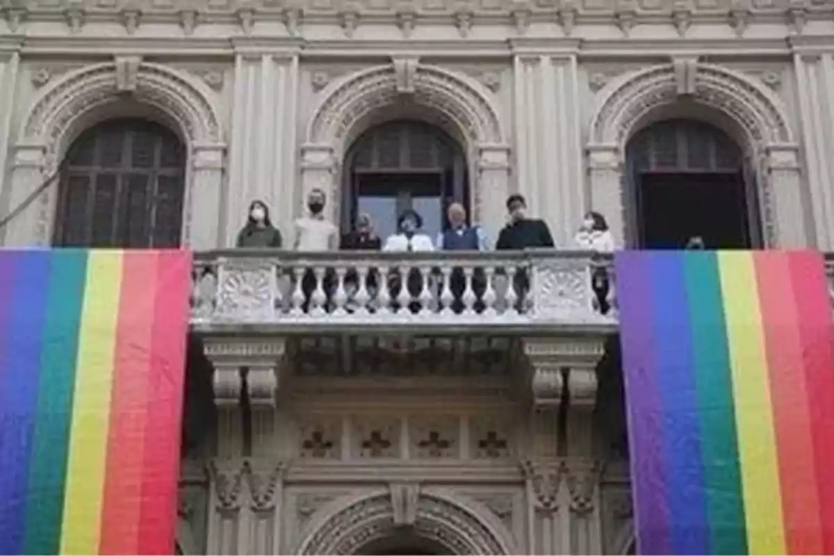 Personas en un balcón de edificio antiguo decorado con dos grandes banderas arcoíris