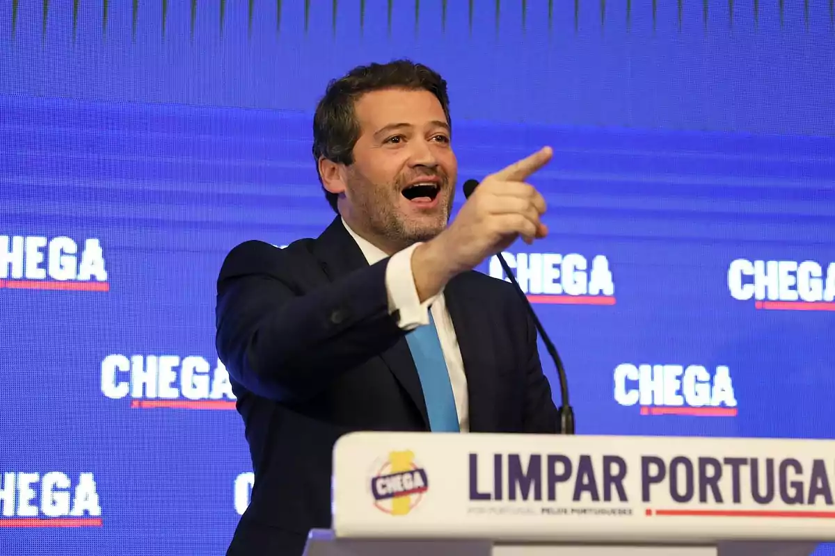 Un hombre en un podio hablando y señalando con el dedo, con un fondo azul que tiene el texto "CHEGA" repetido.