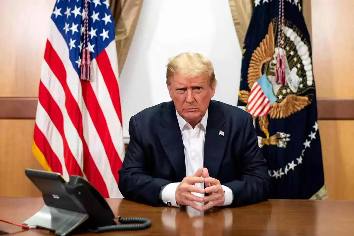 Blond-haired man with a serious expression sitting at a table in front of a phone with United States flags in the background Blond-haired man with a serious expression sitting at a table in front of a phone with United States flags in the background