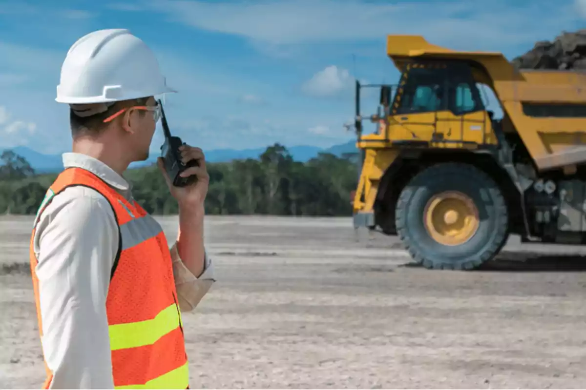 Trabajador de construcción con casco y chaleco de seguridad usando un walkie-talkie frente a un camión volquete en un sitio de construcción.