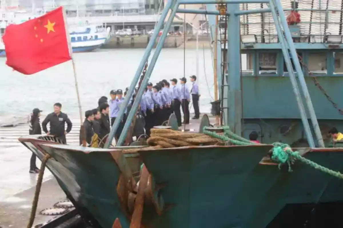 Un grupo de personas uniformadas está de pie en un muelle junto a un barco con una bandera roja ondeando.