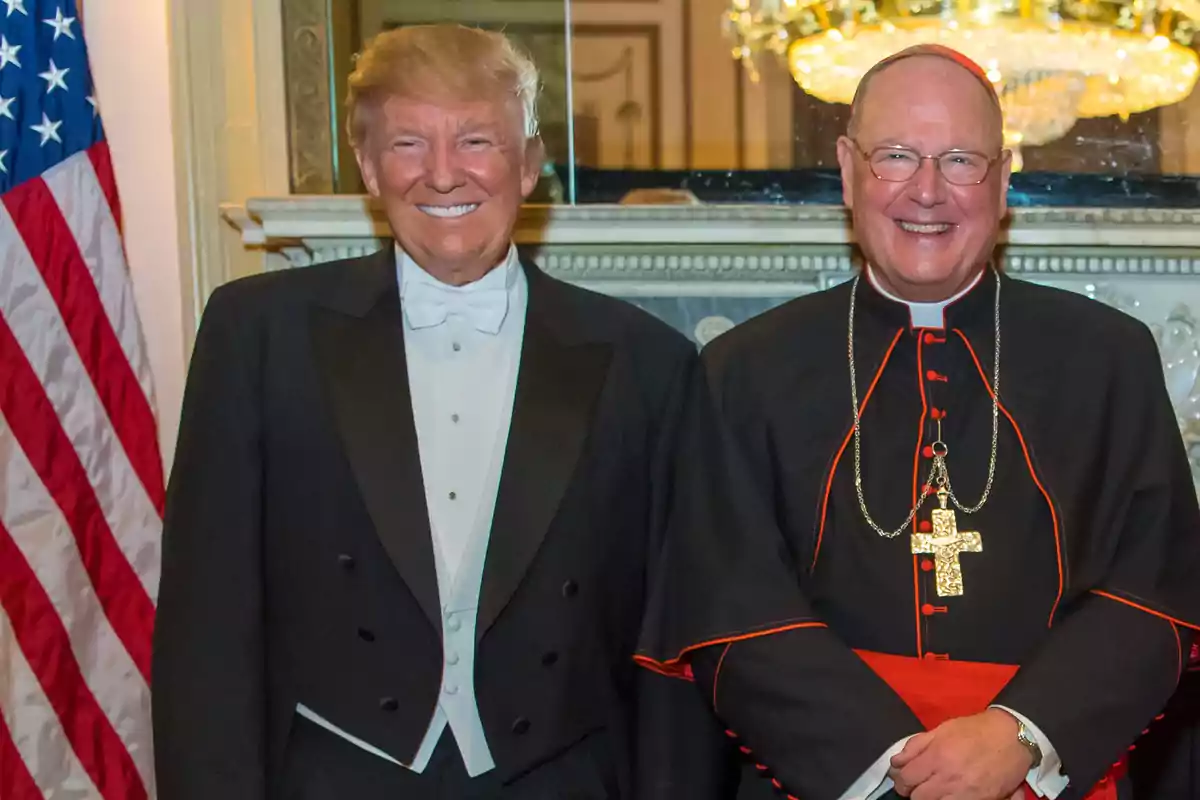 Dos hombres sonrientes posan juntos, uno con esmoquin y el otro con vestimenta religiosa, frente a una bandera y un candelabro. Dos hombres sonrientes posan juntos, uno con esmoquin y el otro con vestimenta religiosa, frente a una bandera y un candelabro.