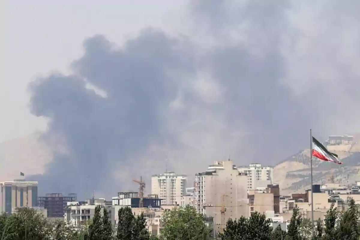 Humo negro se eleva sobre una ciudad con edificios altos y una bandera de Irán ondeando en primer plano Humo negro se eleva sobre una ciudad con edificios altos y una bandera de Irán ondeando en primer plano