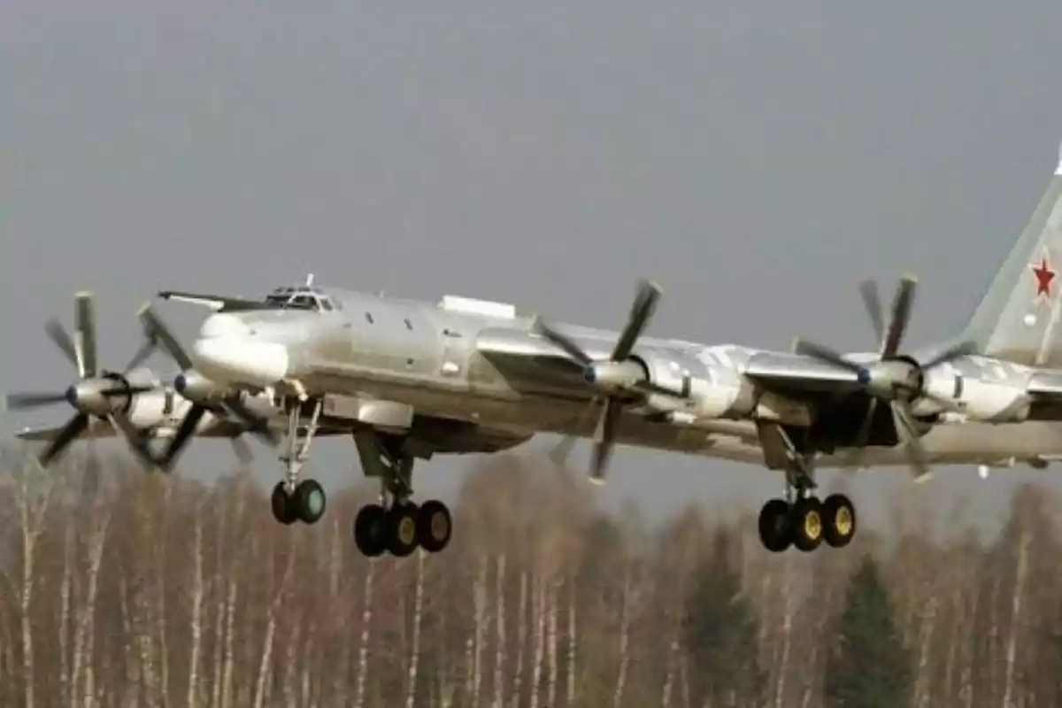Avión militar de hélices en vuelo bajo con tren de aterrizaje desplegado sobre un fondo de árboles y cielo nublado Avión militar de hélices en vuelo bajo con tren de aterrizaje desplegado sobre un fondo de árboles y cielo nublado