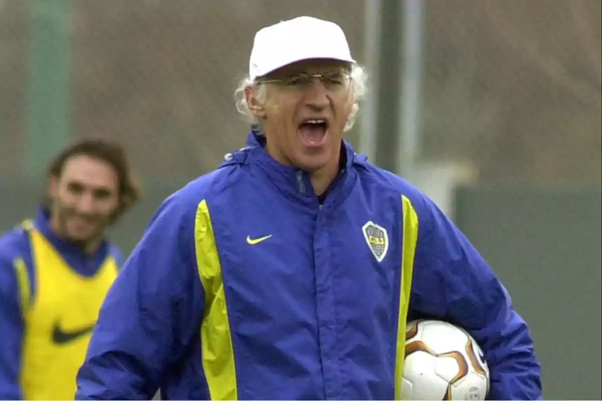 A man in a blue and yellow jacket and a white cap holds a soccer ball while appearing to be talking or shouting. A man in a blue and yellow jacket and a white cap holds a soccer ball while appearing to be talking or shouting.
