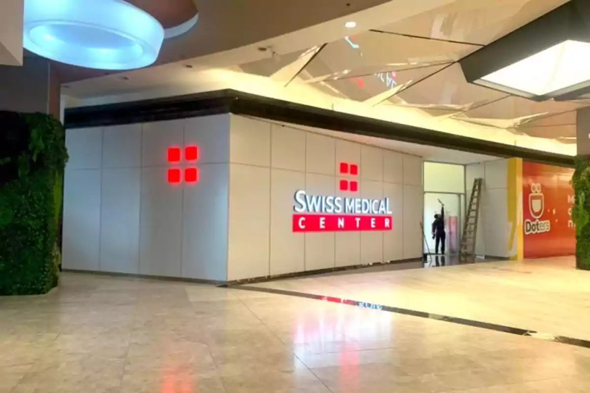 Fachada de un centro médico llamado Swiss Medical Center en el interior de un centro comercial con paredes blancas y logotipo iluminado en rojo Fachada de un centro médico llamado Swiss Medical Center en el interior de un centro comercial con paredes blancas y logotipo iluminado en rojo