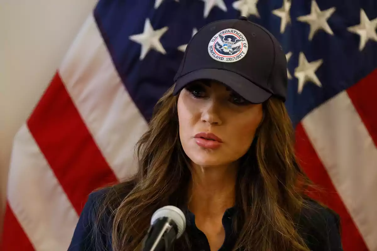 Woman wearing a Department of Homeland Security cap speaking into a microphone with a United States flag in the background