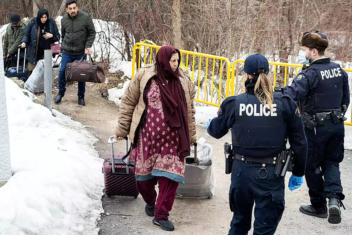 Personas con equipaje caminando por un camino nevado mientras son observadas por oficiales de policía.