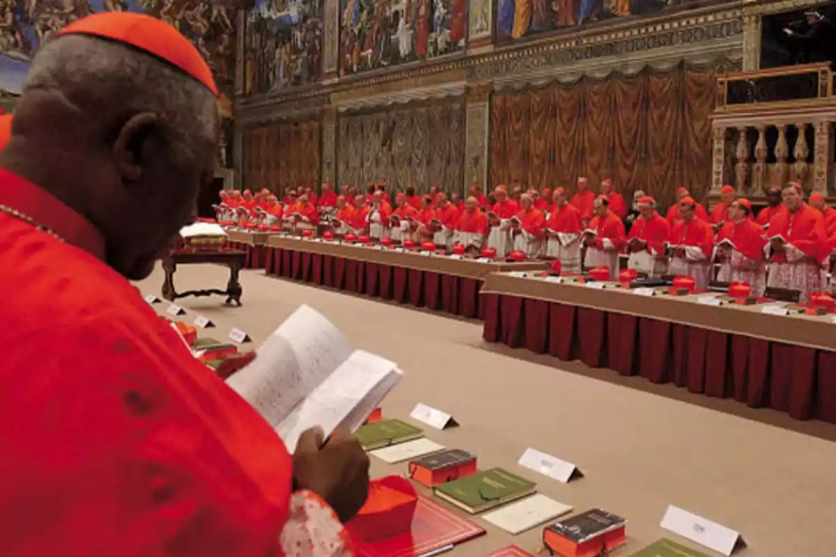 Un grupo de cardenales vestidos de rojo se reúne en una sala decorada con frescos, mientras uno de ellos lee un documento.