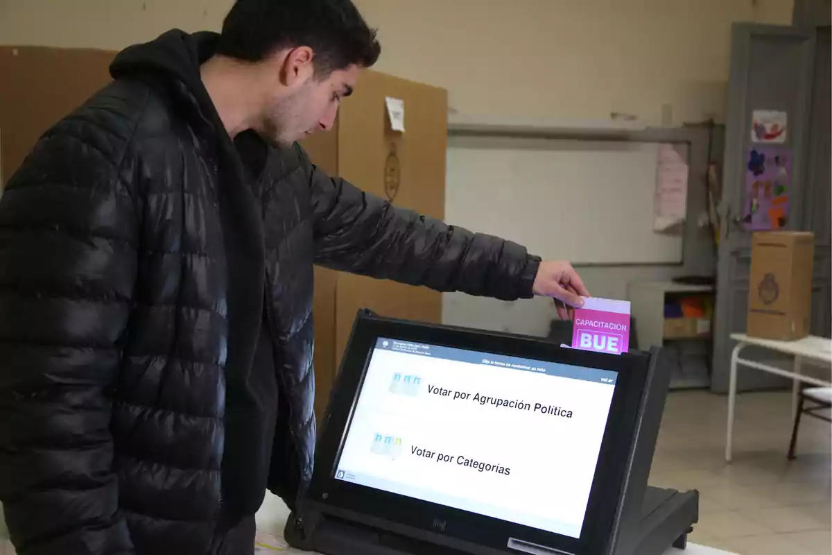 Un hombre con chaqueta negra está utilizando una máquina de votación electrónica en un centro de votación, insertando una tarjeta de capacitación en la ranura de la máquina.