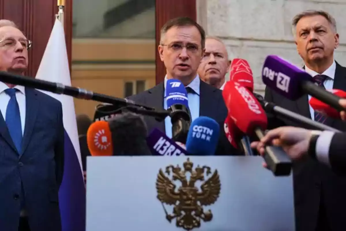 A group of men in suits is standing in front of several microphones from different media outlets, with an official emblem visible on the podium.