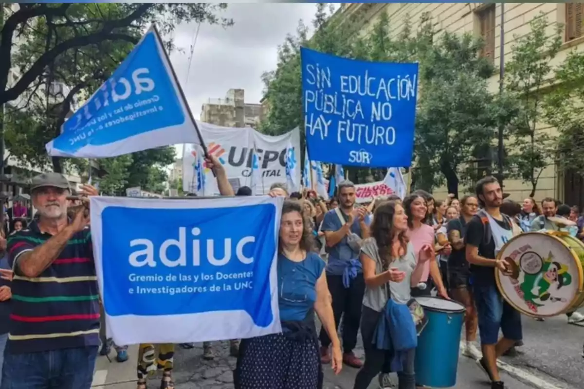 Personas marchando por la calle con pancartas y banderas en defensa de la educación pública, algunas personas tocan tambores y otras sostienen carteles azules Personas marchando por la calle con pancartas y banderas en defensa de la educación pública, algunas personas tocan tambores y otras sostienen carteles azules