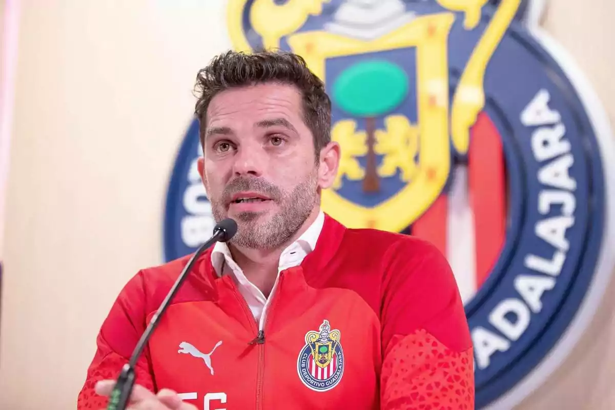 Hombre con chaqueta roja de Chivas hablando en una conferencia de prensa con el escudo del club de fondo Hombre con chaqueta roja de Chivas hablando en una conferencia de prensa con el escudo del club de fondo