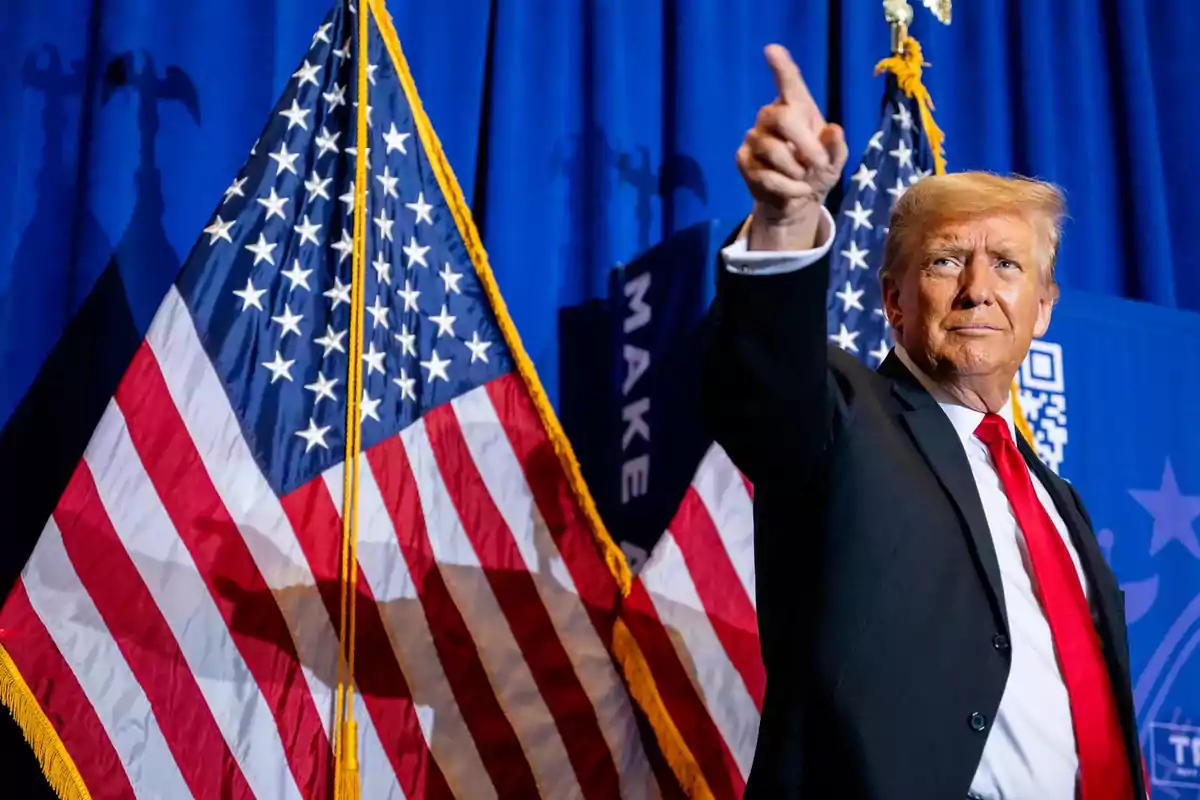 Man in a suit with a red tie pointing upward in front of two American flags and a blue background