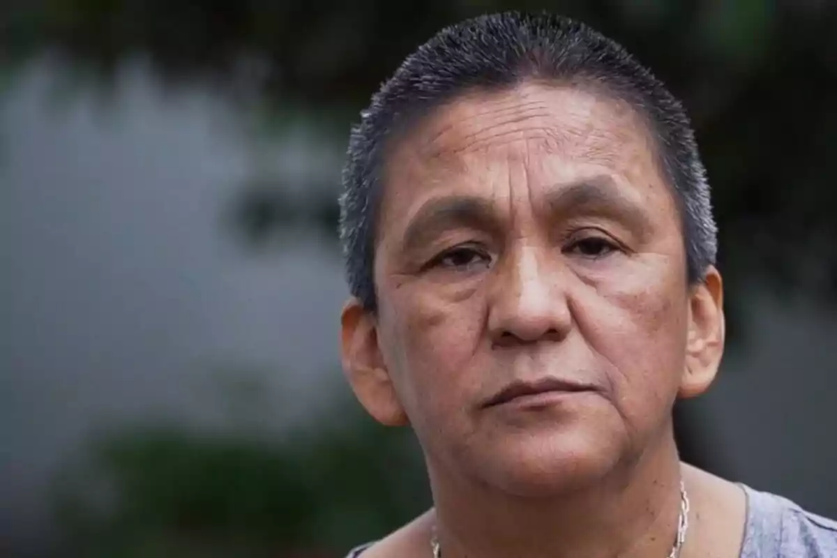 Una persona con expresión seria y cabello corto en un entorno exterior. Una persona con expresión seria y cabello corto en un entorno exterior.