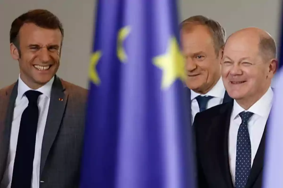 Tres hombres sonrientes detrás de una bandera de la Unión Europea.