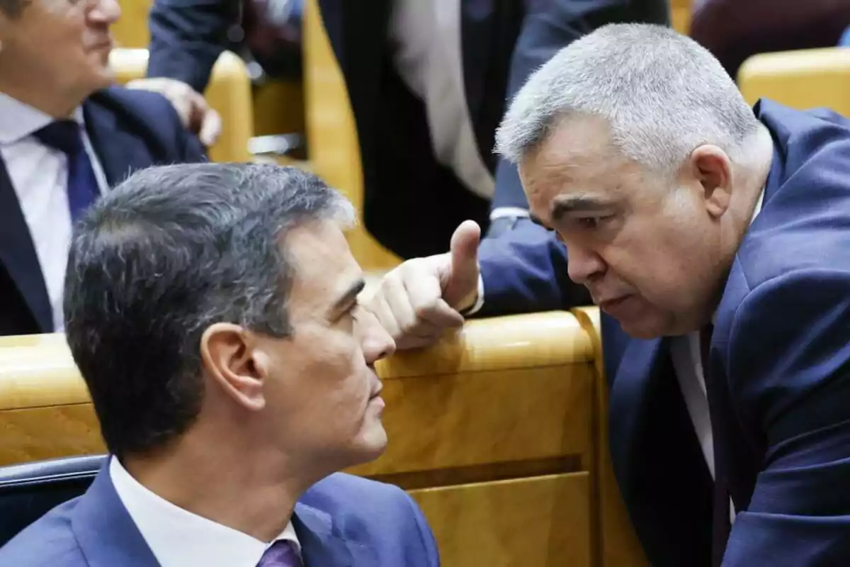 Two men in suits are conversing closely in a formal setting, possibly in a parliament or meeting room.