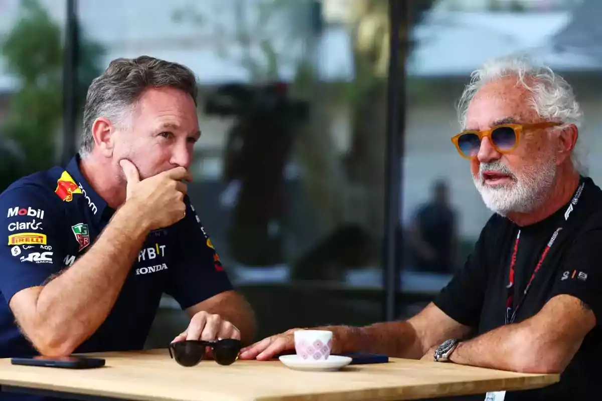 Dos hombres conversan sentados en una mesa de madera al aire libre uno lleva una camiseta de equipo de automovilismo y el otro usa gafas de sol y tiene barba blanca sobre la mesa hay una taza de café y unos lentes oscuros Dos hombres conversan sentados en una mesa de madera al aire libre uno lleva una camiseta de equipo de automovilismo y el otro usa gafas de sol y tiene barba blanca sobre la mesa hay una taza de café y unos lentes oscuros