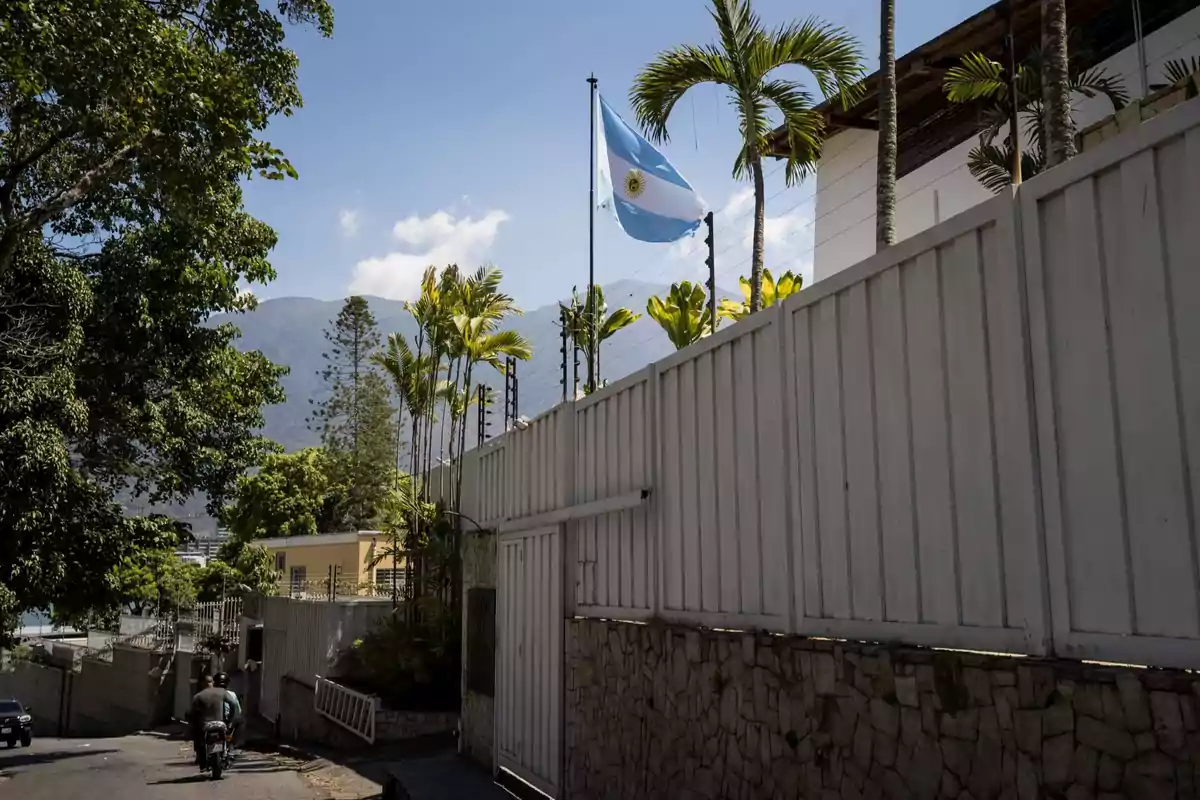 Una bandera ondea en un mástil junto a un edificio rodeado de palmeras y un muro blanco, con montañas al fondo y un cielo despejado.