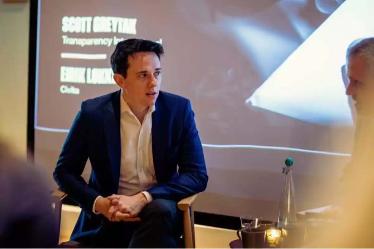 A man sitting in a chair during a presentation or conference, with a background showing text on a screen.