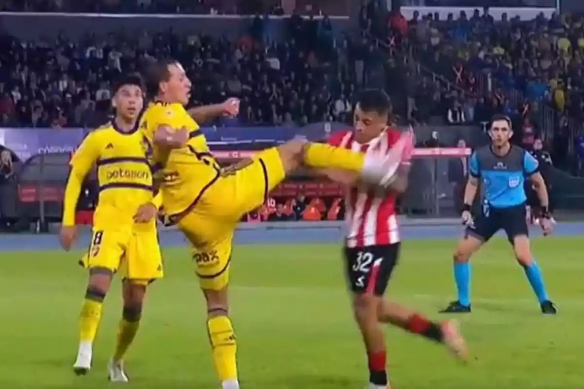 Un jugador de fútbol con uniforme amarillo levanta la pierna en un intento de despejar el balón mientras un oponente con uniforme a rayas rojas y blancas se protege.