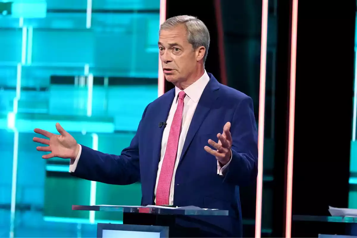 Man in a blue suit and pink tie gesturing on a stage with a blue background.