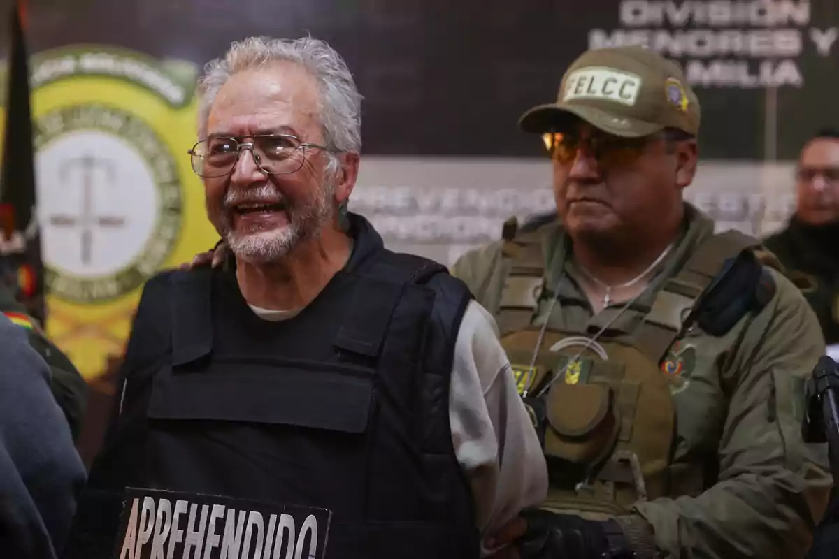 An elderly man wearing a bulletproof vest and glasses is escorted by a police officer in uniform and cap in what appears to be a press conference or official presentation setting.