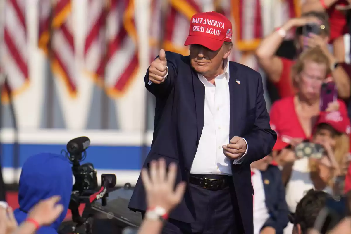 Un hombre con gorra roja hace un gesto de aprobación con el pulgar levantado en un evento al aire libre con banderas de fondo.