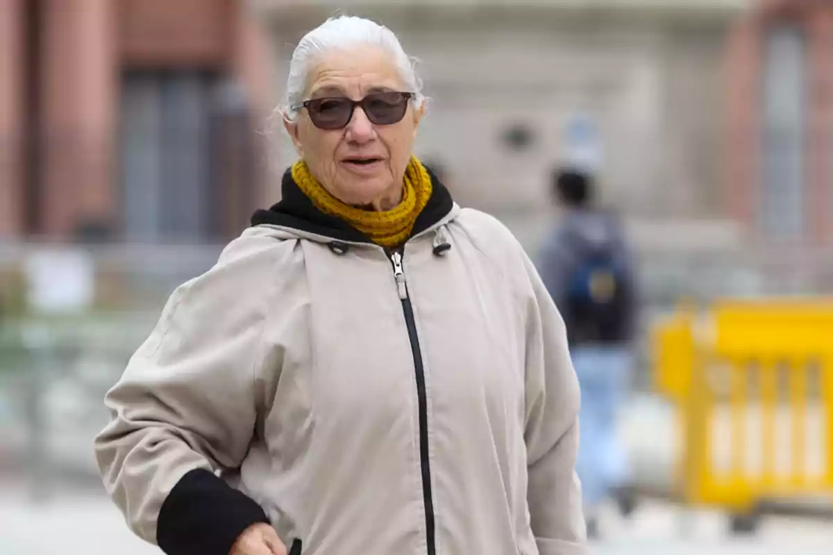 Una mujer mayor con gafas de sol y bufanda amarilla lleva una chaqueta clara mientras camina al aire libre.