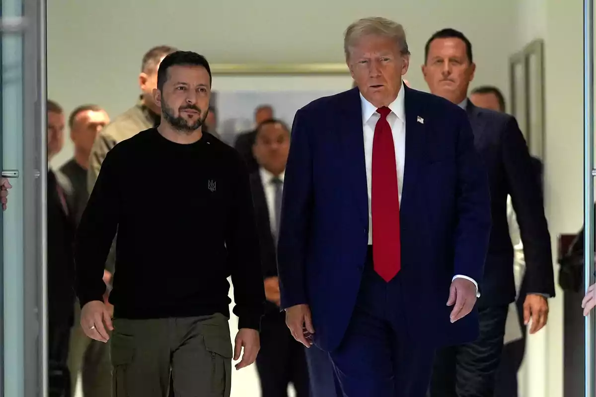 Un grupo de personas caminando por un pasillo, con dos hombres al frente, uno con traje azul y corbata roja y el otro con suéter negro y pantalones verdes.