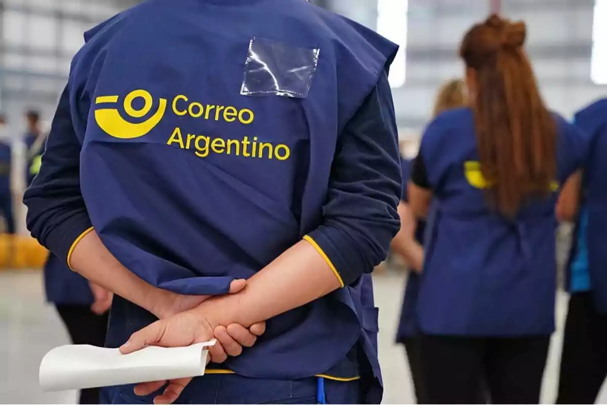 Una persona con uniforme azul del Correo Argentino sostiene un papel en la mano mientras está de pie en un entorno de trabajo con otras personas al fondo.