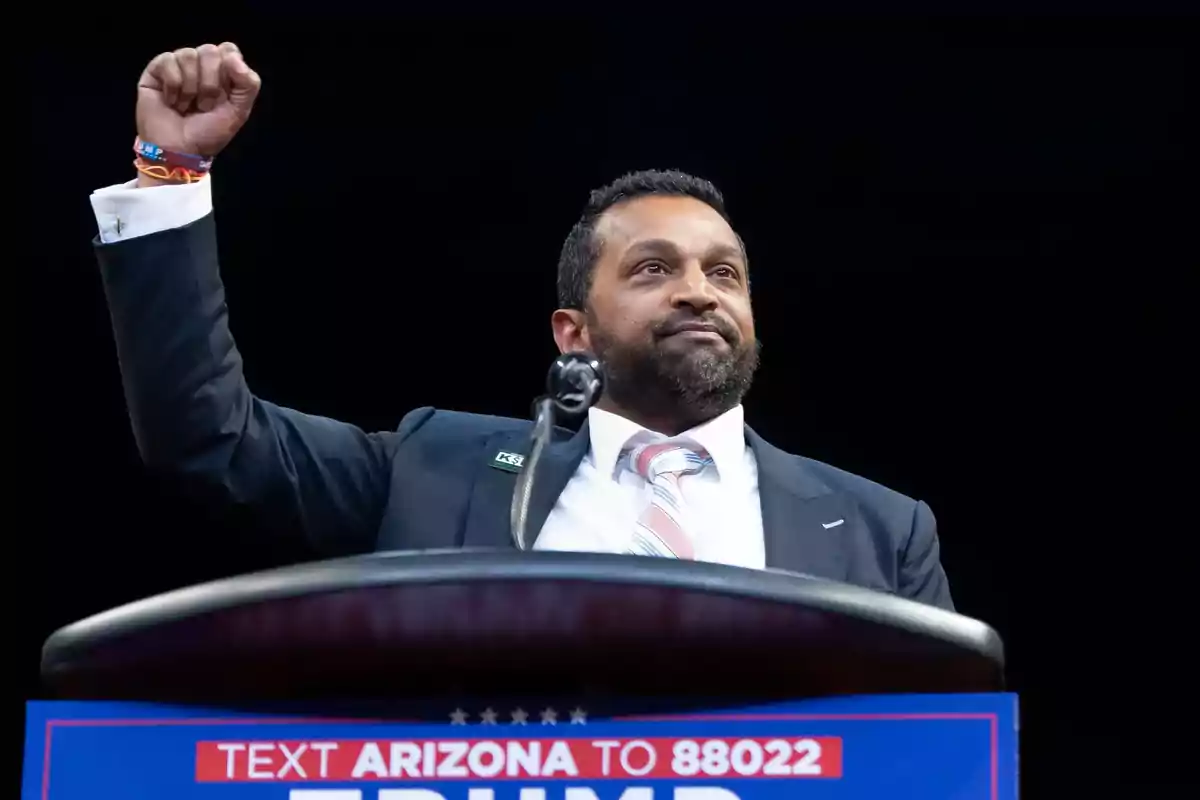 Hombre en un podio levantando el puño con un micrófono frente a él y un cartel que dice "TEXT ARIZONA TO 88022 TRUMP".