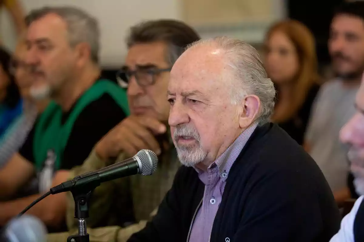 Un hombre mayor hablando en un micrófono durante una reunión con varias personas sentadas a su alrededor.