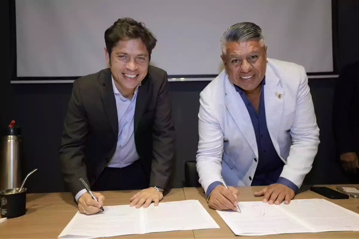 Dos hombres sonrientes firmando documentos en una mesa de madera con una pantalla de fondo.