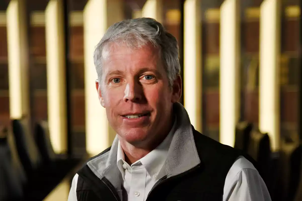 A gray-haired man with light eyes wearing a dark jacket and a white jersey in a well-lit indoor setting. A gray-haired man with light eyes wearing a dark jacket and a white jersey in a well-lit indoor setting.