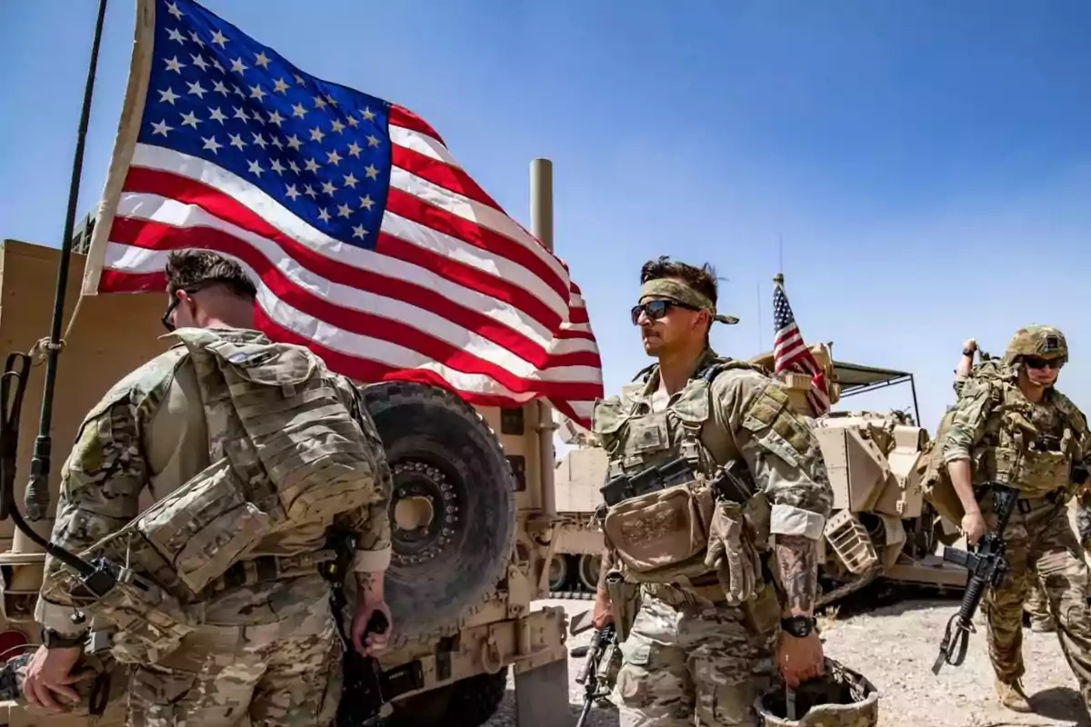 Soldados en uniforme militar caminan junto a un vehículo con una bandera de Estados Unidos ondeando al fondo.