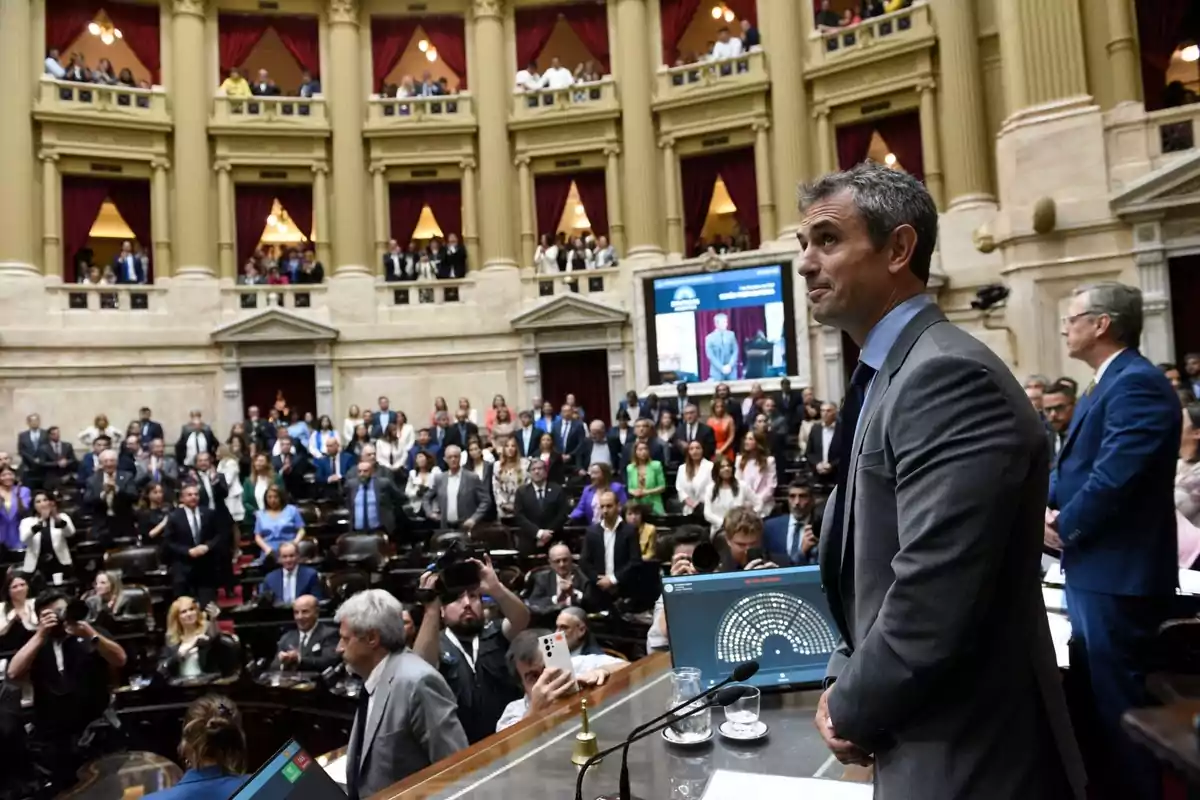 Una persona de pie en un recinto legislativo lleno de gente, con balcones y cortinas rojas en el fondo.