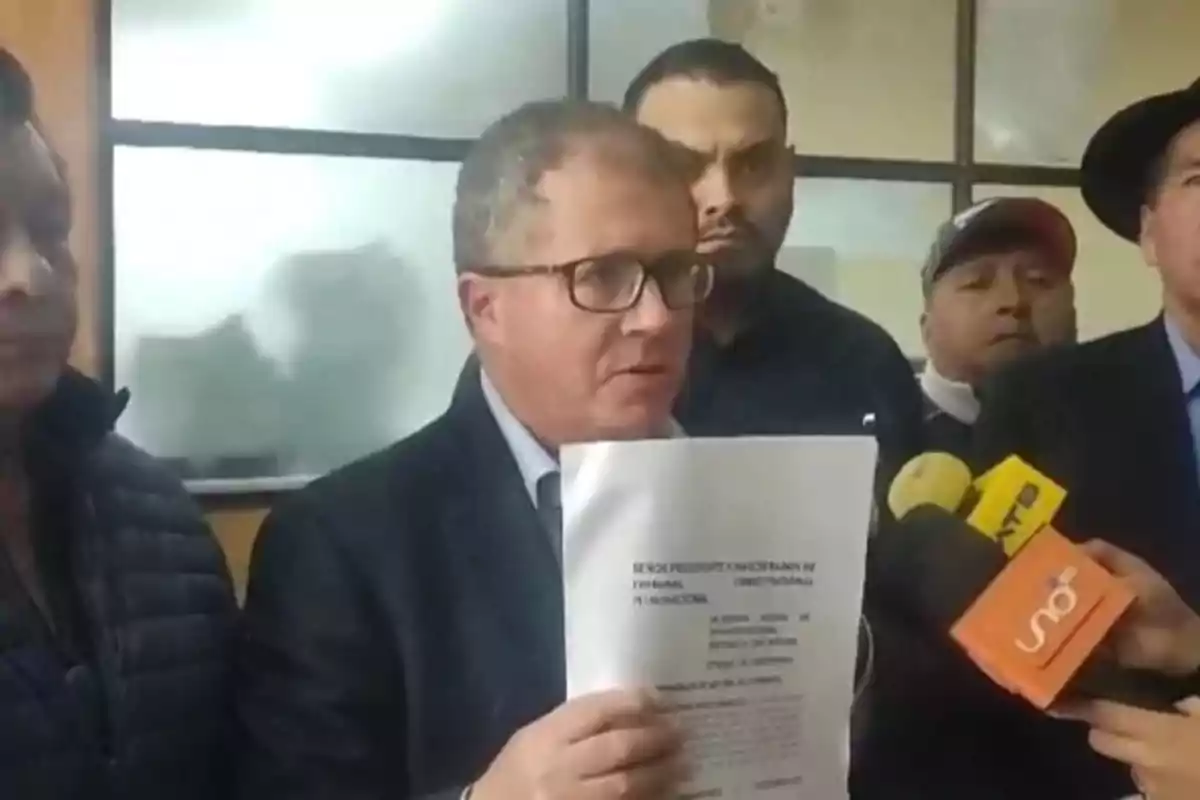 Un hombre con gafas sostiene un documento mientras habla frente a varios micrófonos y está rodeado de otras personas en una sala.