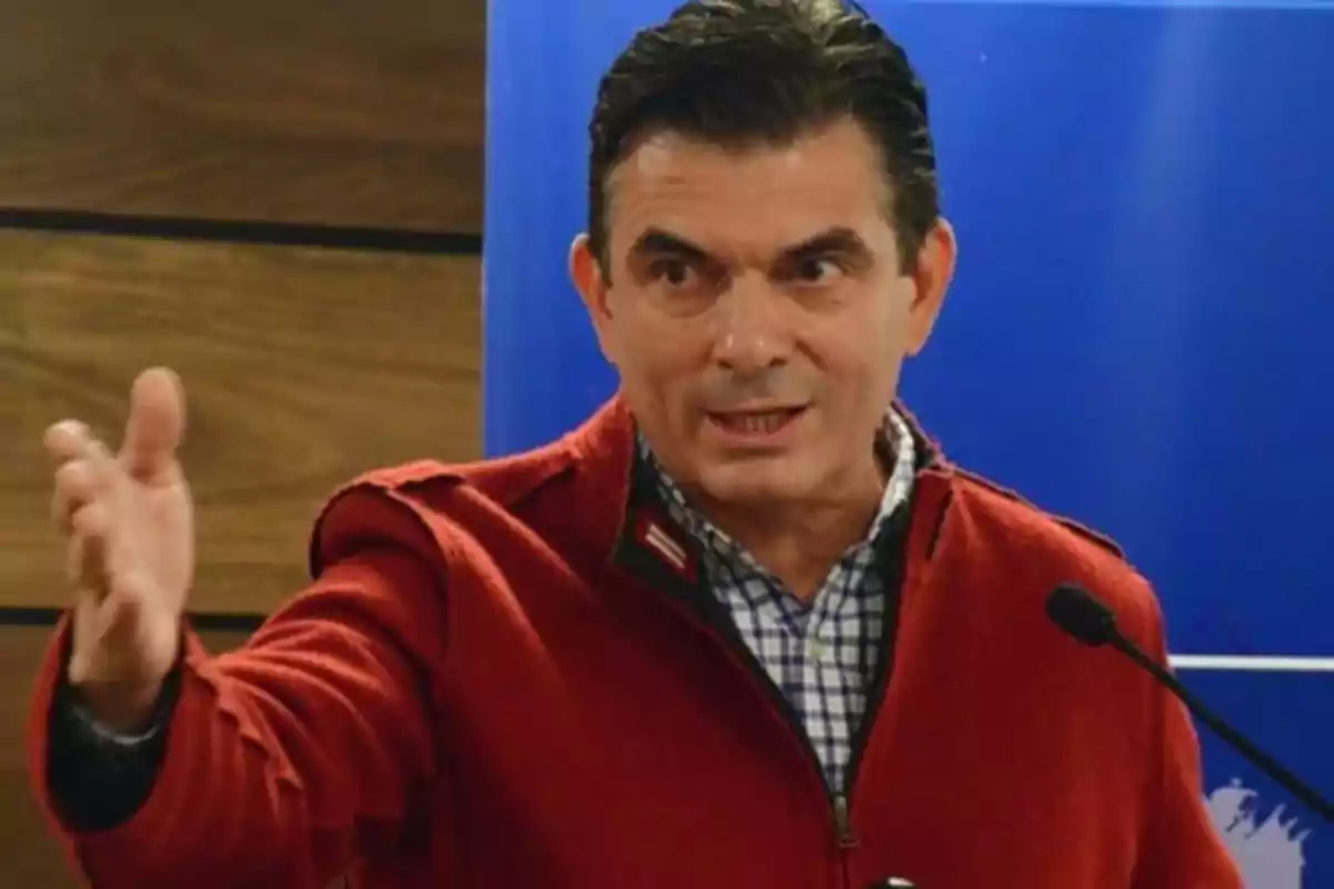 A man in a red jacket gestures while speaking in front of a microphone.