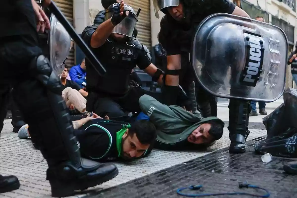 Policías antidisturbios detienen a dos personas en el suelo durante una manifestación en la calle.