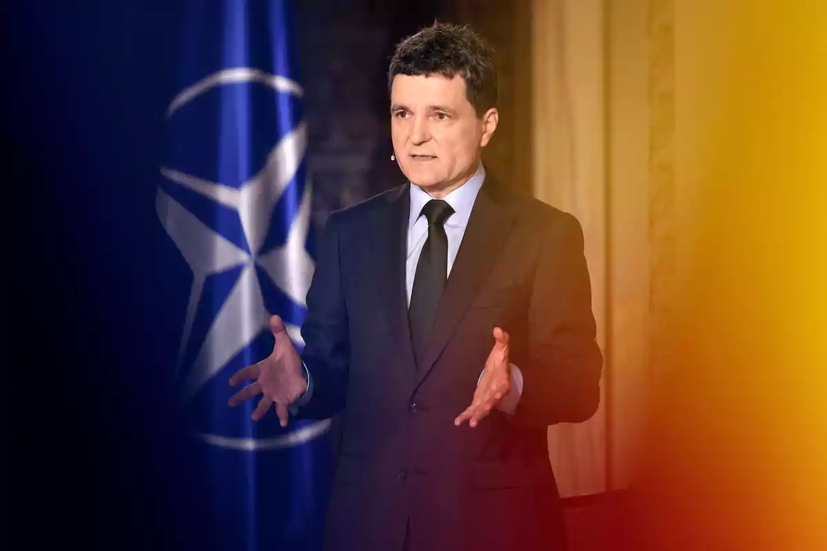 A man in a dark suit and black tie gestures while speaking, with a blue flag featuring a white star symbol in the background. A man in a dark suit and black tie gestures while speaking, with a blue flag featuring a white star symbol in the background.