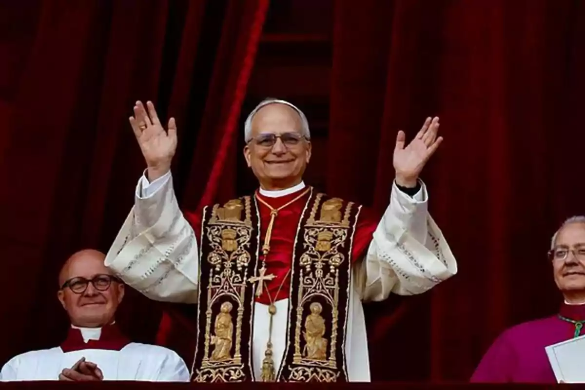 Un hombre con vestimenta religiosa levanta las manos y sonríe mientras está de pie entre dos personas frente a un fondo de cortinas rojas Un hombre con vestimenta religiosa levanta las manos y sonríe mientras está de pie entre dos personas frente a un fondo de cortinas rojas
