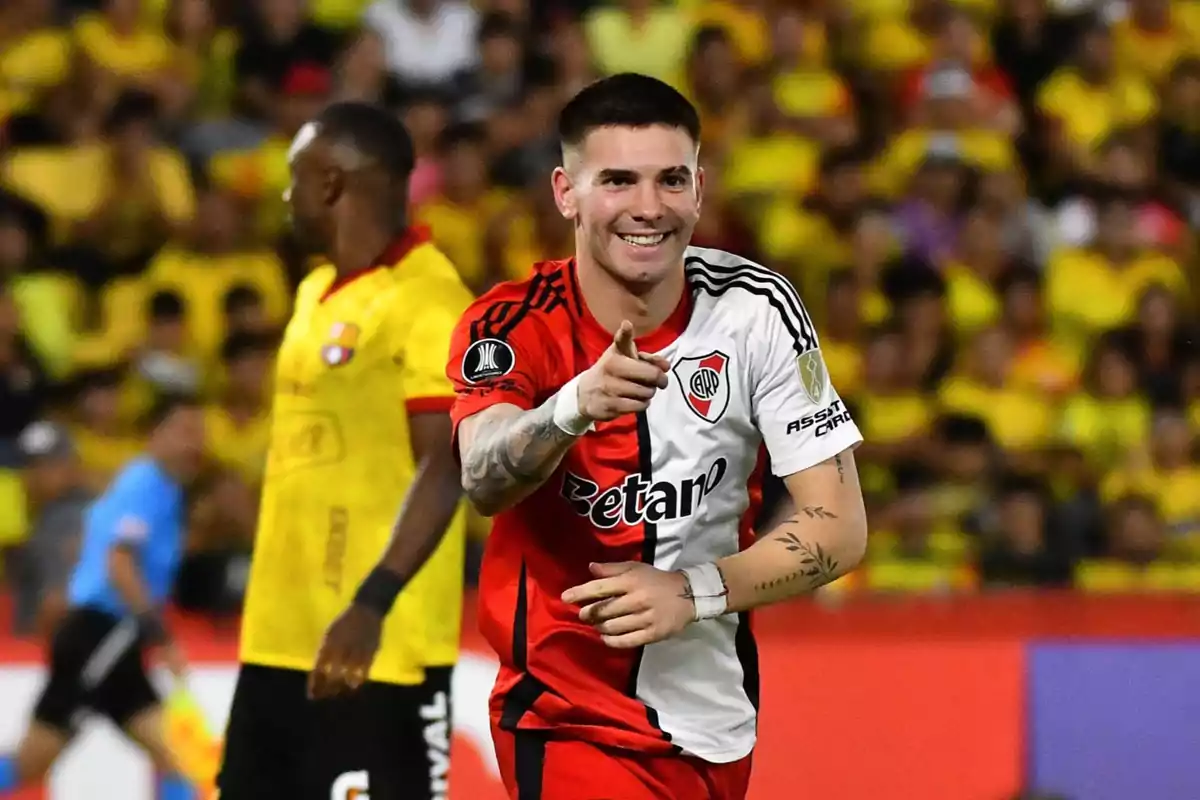 Un jugador de fútbol con uniforme rojo y blanco sonríe y señala hacia adelante en un estadio lleno de espectadores. Un jugador de fútbol con uniforme rojo y blanco sonríe y señala hacia adelante en un estadio lleno de espectadores.