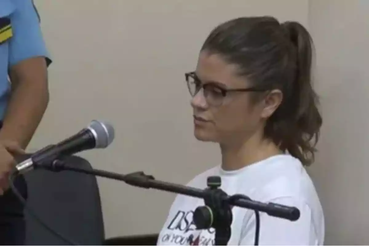 Mujer con gafas y cabello recogido sentada frente a un micrófono en una sala junto a una persona con uniforme azul Mujer con gafas y cabello recogido sentada frente a un micrófono en una sala junto a una persona con uniforme azul