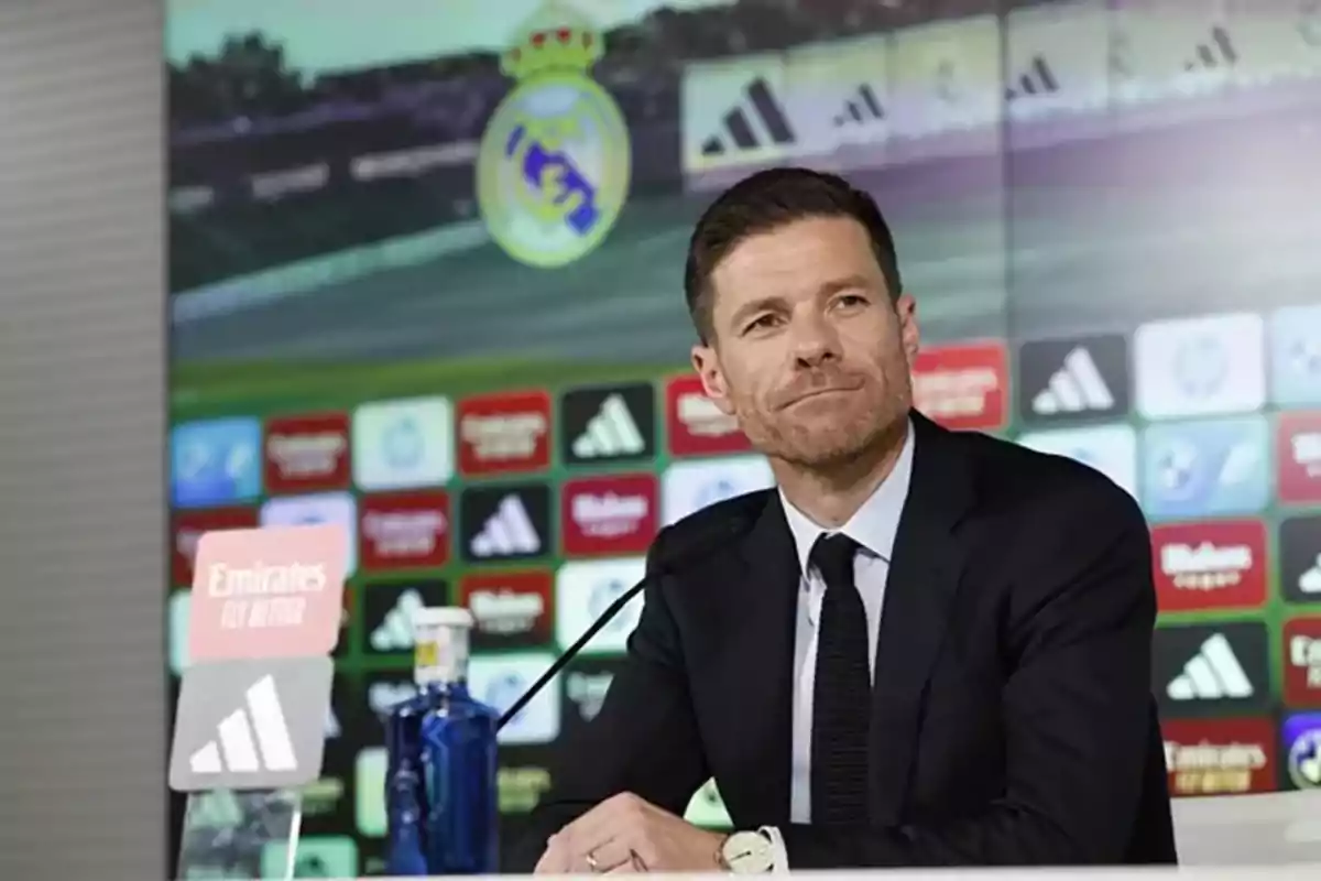 Hombre con traje oscuro sentado en una conferencia de prensa con el escudo del Real Madrid y varios logotipos de patrocinadores al fondo Hombre con traje oscuro sentado en una conferencia de prensa con el escudo del Real Madrid y varios logotipos de patrocinadores al fondo